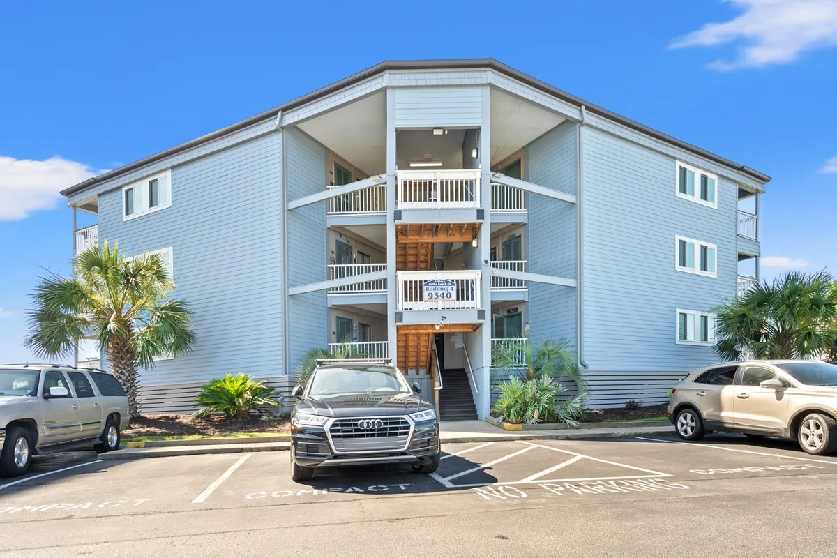 This image captures the exterior view of a three-story condo building with palm trees, a welcoming front staircase, and parked cars, all under sunny skies. The setting suggests an inviting residential environment ideal for longer-term stays, with outdoor areas perfect for relaxation or casual gatherings.