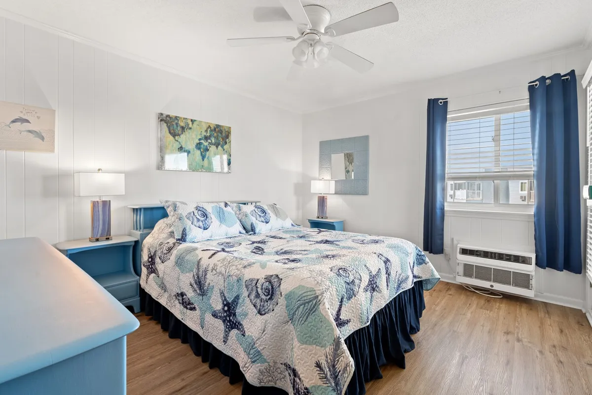 A tranquil primary bedroom with a plush bed dressed in a blue and white quilt, flanked by matching nightstands and lamps. Ocean-themed artwork above the bed and a large window with curtains fill the room with natural morning light, creating a serene retreat for relaxation.