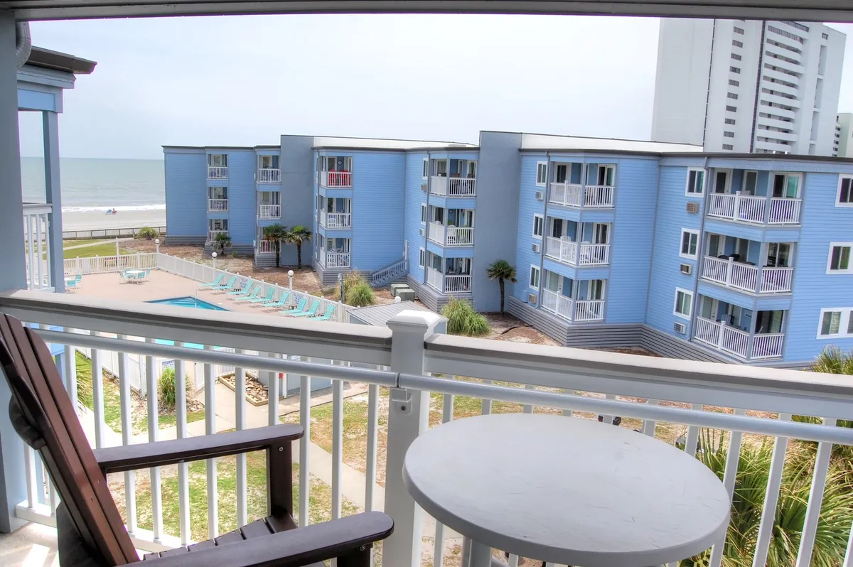 A stunning view from a balcony looking out over a sparkling pool area, with the ocean and resort grounds in the background. The scene blends balcony comfort with lively resort amenities just steps away, including the inviting pool and sun decks.