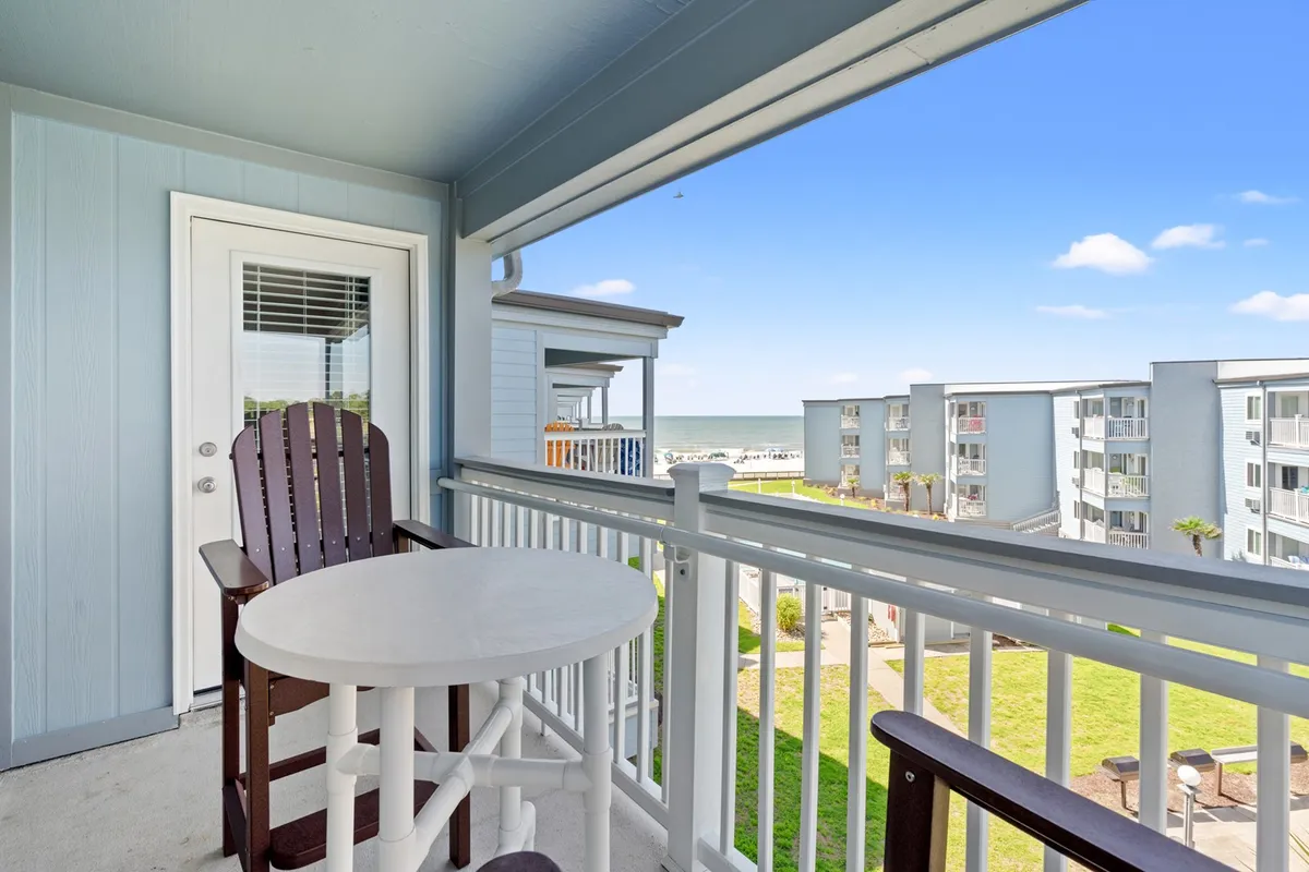 The image showcases the condo's private balcony furnished with cozy seating, overlooking a breathtaking ocean vista. The architecture of the building frames the sweeping sea view, inviting guests to relax and soak in the calming atmosphere. Hints of a vibrant sunset and the gentle sounds of the waves evoke a serene, beachside retreat.