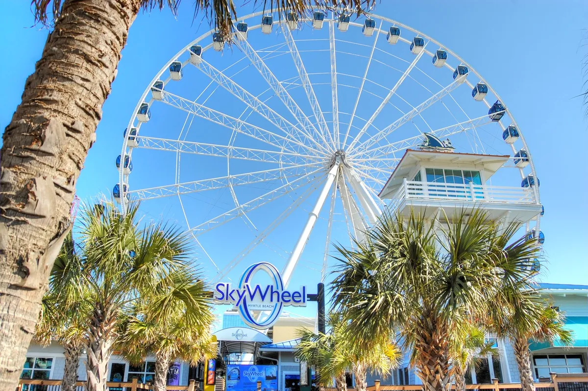 A lively shot of the iconic Myrtle Beach SkyWheel framed by palm trees and a welcoming entrance sign. Visitors are seen gathering and enjoying the vibrant atmosphere, ready to experience panoramic ocean views from above.