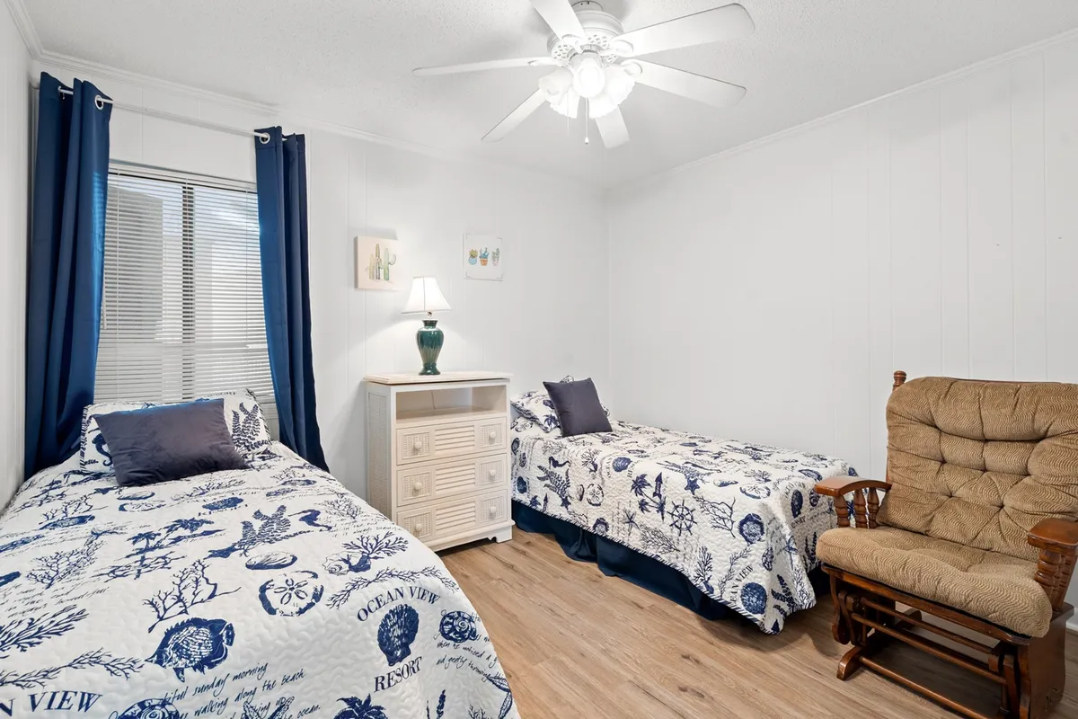 A cozy second bedroom featuring two twin beds, a nightstand with a lamp between them, and a comfortable rocking chair. The setting is calm and inviting, perfect for children or friends to unwind after a day of adventures.