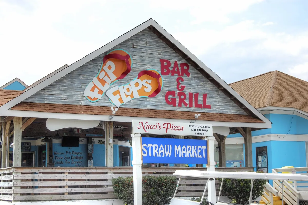 A busy corner featuring the Flip Flops Bar & Grill, Nucci’s Pizza, and Straw Market signage. The area is lively with visitors dining, shopping, and mingling in a relaxed, cheerful environment.