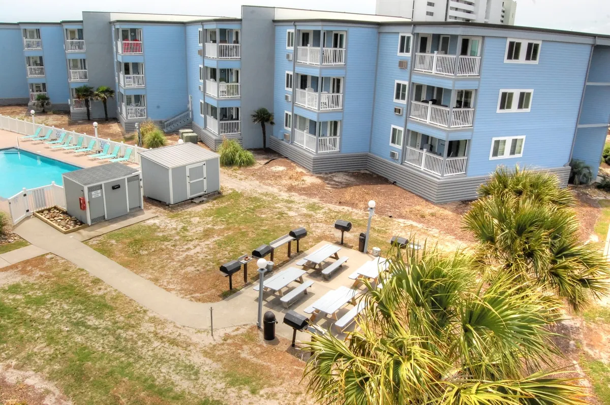 A bright outdoor scene showcasing a sunny swimming pool area with a barbecue station, surrounded by well-kept landscaping and adjacent buildings. Guests are shown mingling, swimming, and grilling, capturing the lively, social vibe of the resort.