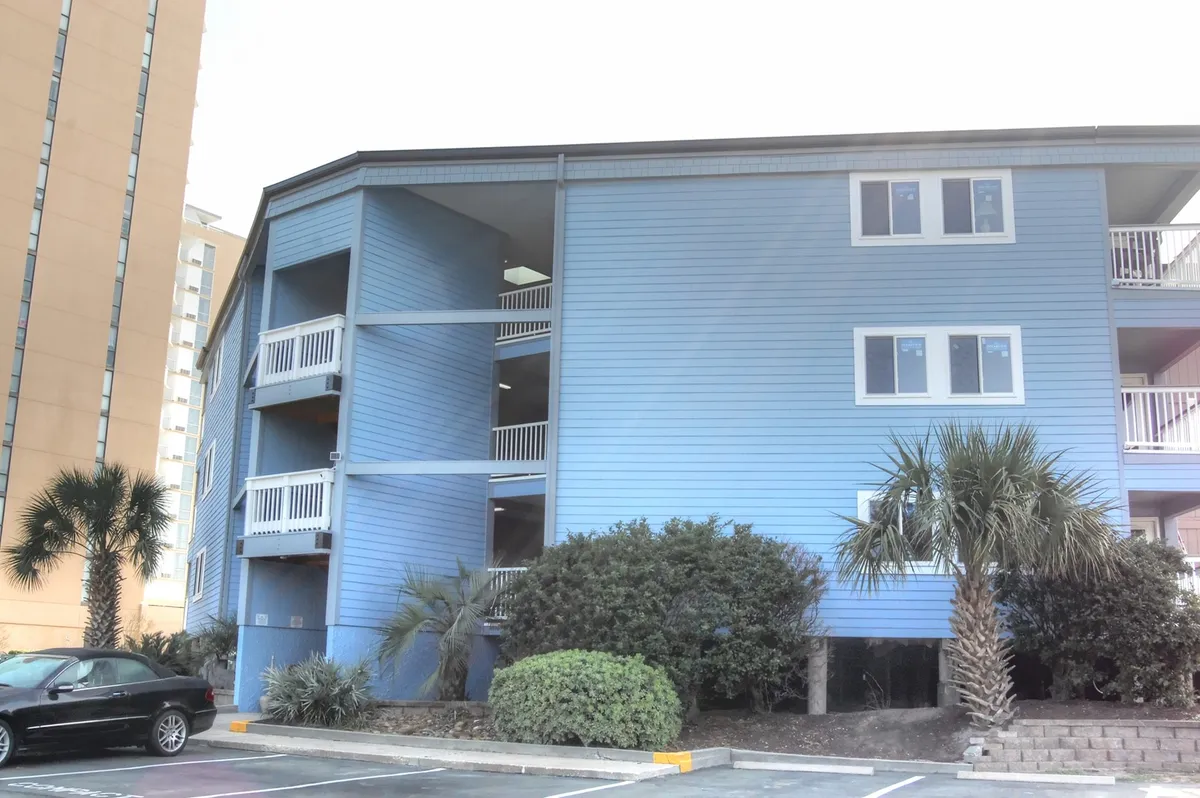 The blue exterior of a vacation building with palm trees and balconies. People can be seen relaxing outdoors, enjoying the garden or socializing, set against a cheerful backdrop under sunny skies.