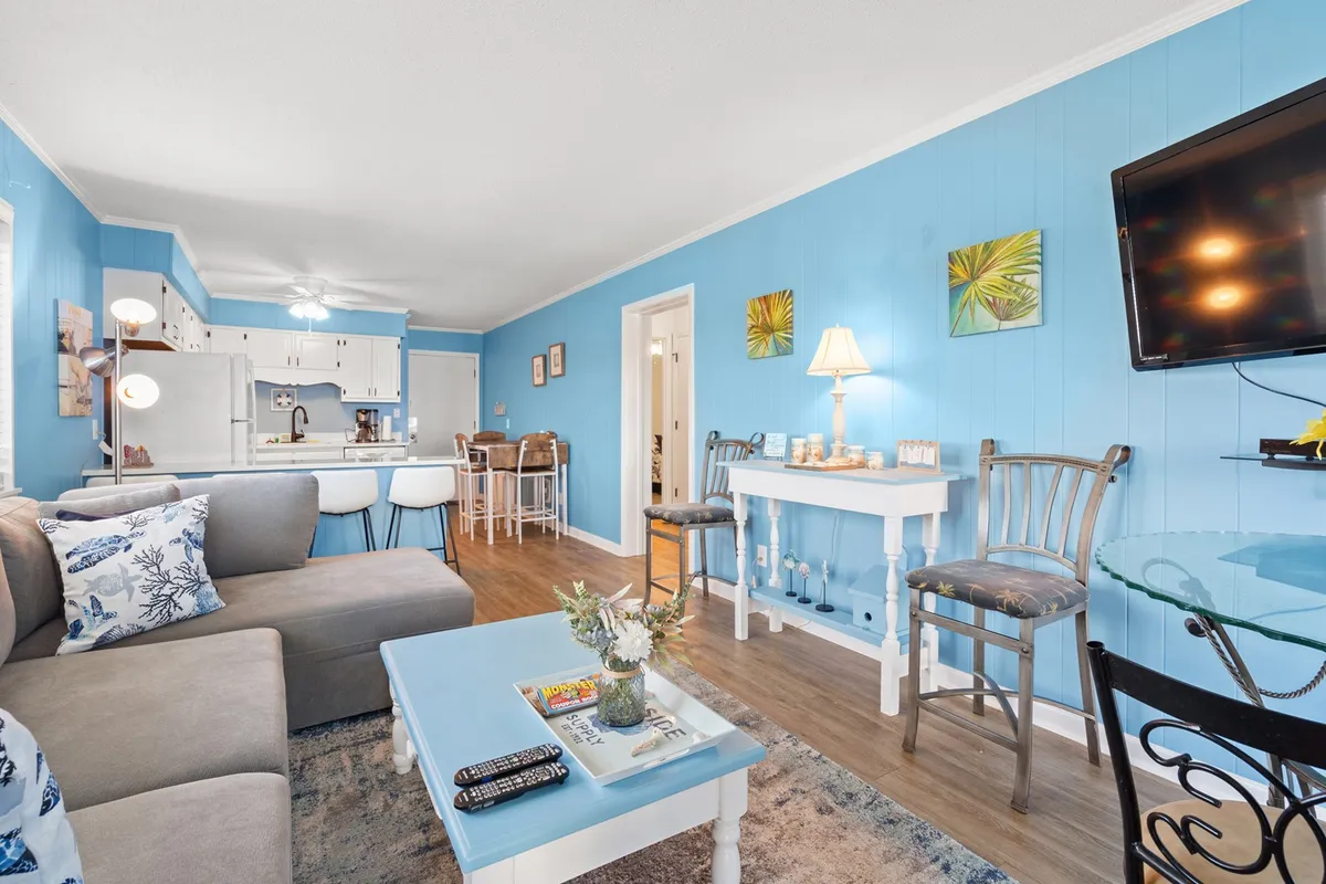 This image depicts a casual, inviting living space with a plush sofa, light blue walls, a coffee table, decorative accents on a side table, and a view into the dining area. The space is filled with natural light, giving it a bright and uplifting atmosphere. The layout encourages socializing and relaxation, making it ideal for gatherings and leisure.