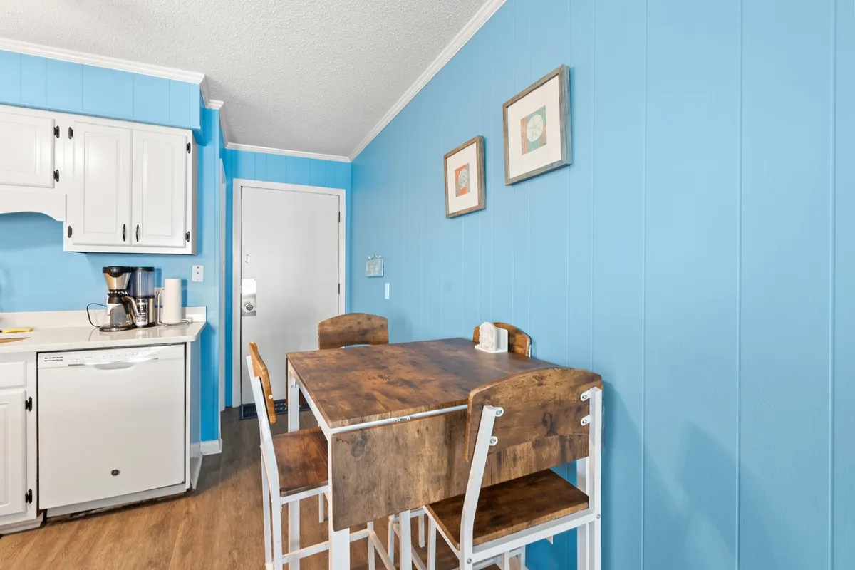 A welcoming dining area featuring a wooden table, blue accent wall, and framed artwork. The space is cozy and designed for shared meals or coffee breaks, with a relaxed and inviting mood perfect for family or group gatherings.