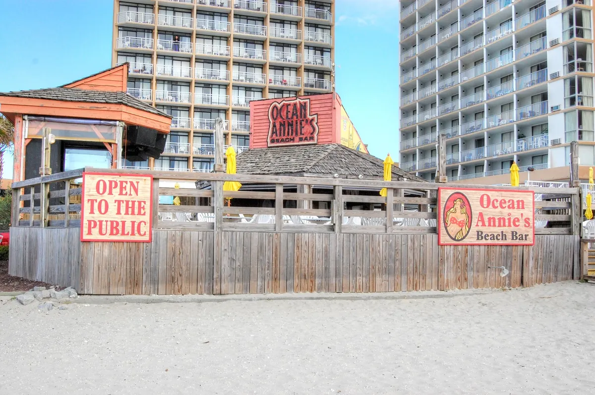 The iconic Ocean Annie's Beach Bar sign is visible above a welcoming wooden deck area, with an 'open to the public' sign inviting guests. The setting hints at the lively yet laid-back ambiance, ideal for evening drinks, live music, and socializing by the ocean.