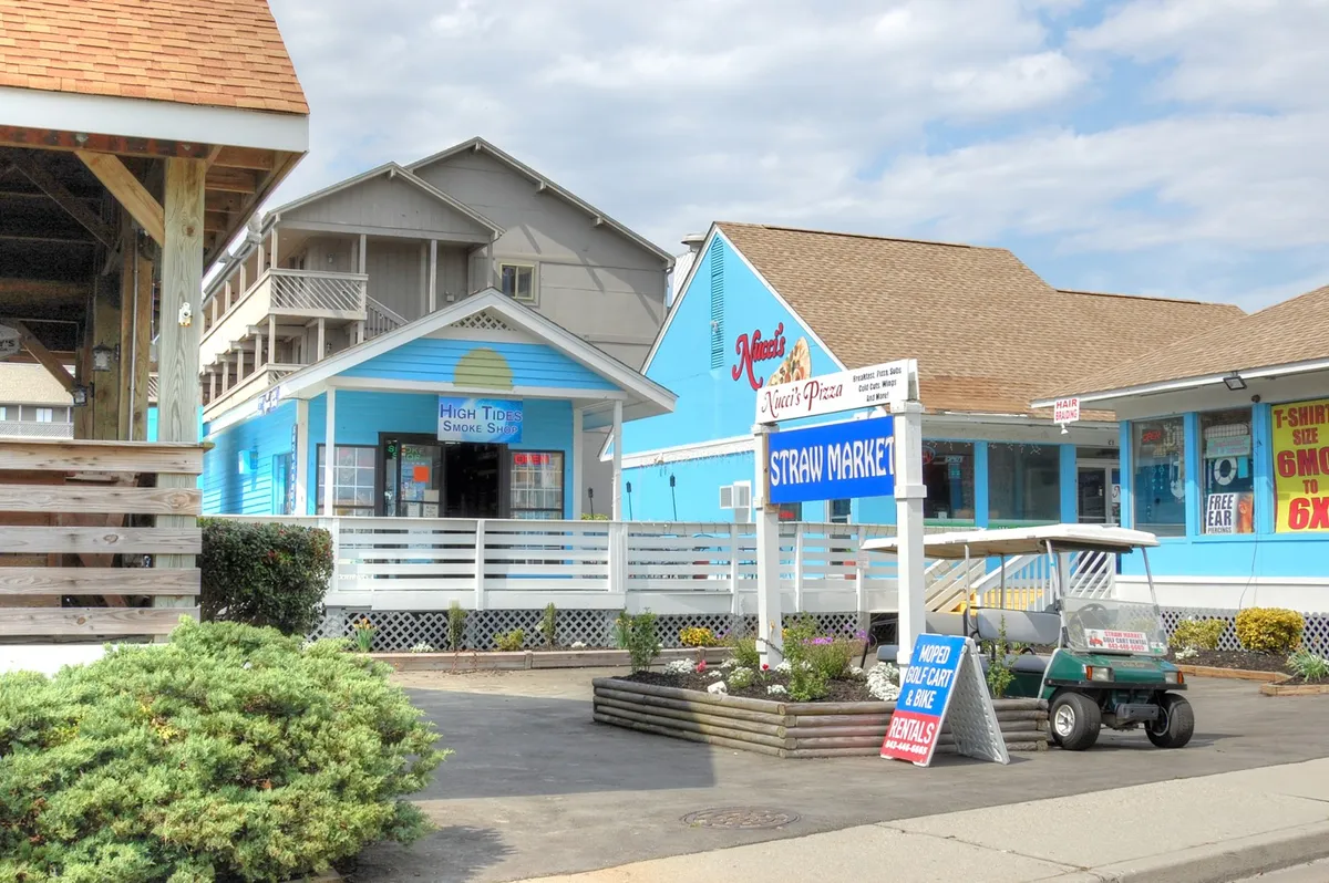 This lively street scene features colorful storefronts, signage for the Straw Market and a pizza shop, and a golf cart in the foreground. The atmosphere is casual and cheerful, representing the convenient access to local dining, shopping, and the laid-back lifestyle that makes long-term stays enjoyable.